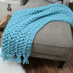 Pu&ograve; includere: Una morbida coperta in crochet blu con frange sul bordo inferiore. La coperta &egrave; drappeggiata su un pouf grigio chiaro.