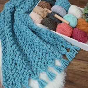 Pu&ograve; includere: Una coperta all'uncinetto blu turchese con frange. La coperta &egrave; realizzata con filato spesso e presenta un motivo a punto testurizzato. La coperta &egrave; appoggiata su un tappeto di pelliccia sintetica bianca.