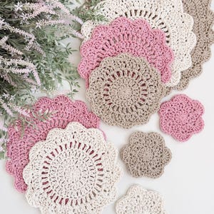 Op de afbeelding: Een verzameling handgemaakte gehaakte doilies en onderzetters in verschillende maten en kleuren. De set bevat ronde doilies in roze, crème en bruin, samen met kleinere, bloemvormige onderzetters. Het haakwerk heeft een geschulpte rand.