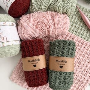 May include: An overhead shot displays a collection of handmade crochet items. There are skeins of yarn in shades of red, green, and pink. Two rolled washcloths, one burgundy and one green, are labeled "Washcloth" and "Dishcloth" with a heart. A crochet hook is also visible.