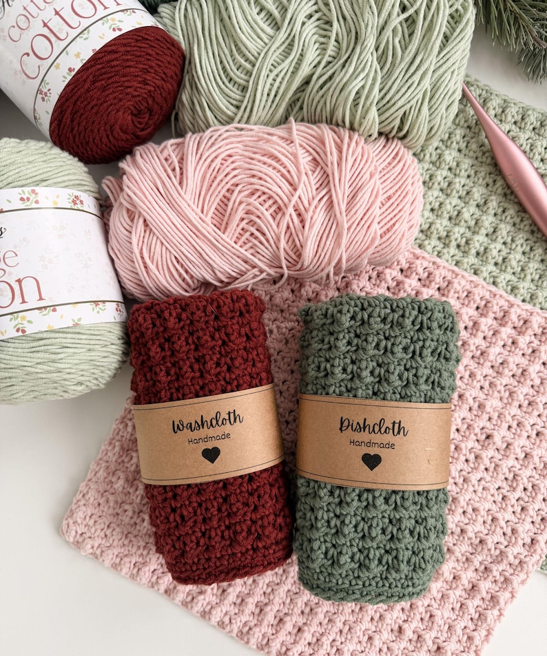 May include: An overhead shot displays a collection of handmade crochet items. There are skeins of yarn in shades of red, green, and pink. Two rolled washcloths, one burgundy and one green, are labeled "Washcloth" and "Dishcloth" with a heart. A crochet hook is also visible.