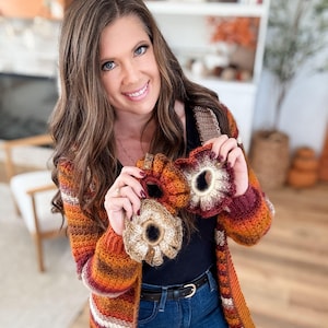 May include: A woman holding three handmade crochet scrunchies in shades of orange, brown, and cream. The scrunchies have a floral design and are accented with small wooden tags. She is wearing a striped orange, brown, and cream cardigan and blue jeans.
