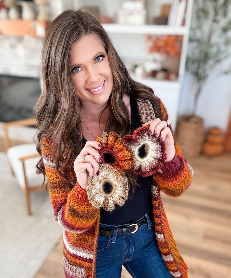 May include: A woman holding three handmade crochet scrunchies in shades of orange, brown, and cream. The scrunchies have a floral design and are accented with small wooden tags. She is wearing a striped orange, brown, and cream cardigan and blue jeans.