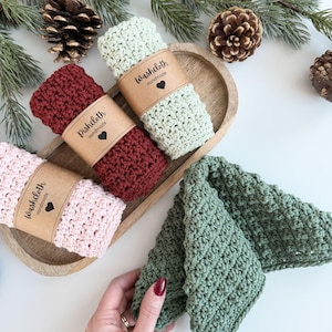 May include: Handmade crochet washcloths in shades of pink, burgundy, and sage green. Each cloth is rolled and secured with a brown paper band, labelled "Washcloth Handmade" with a heart. The washcloths are presented on a wooden tray, with pine cones and foliage.