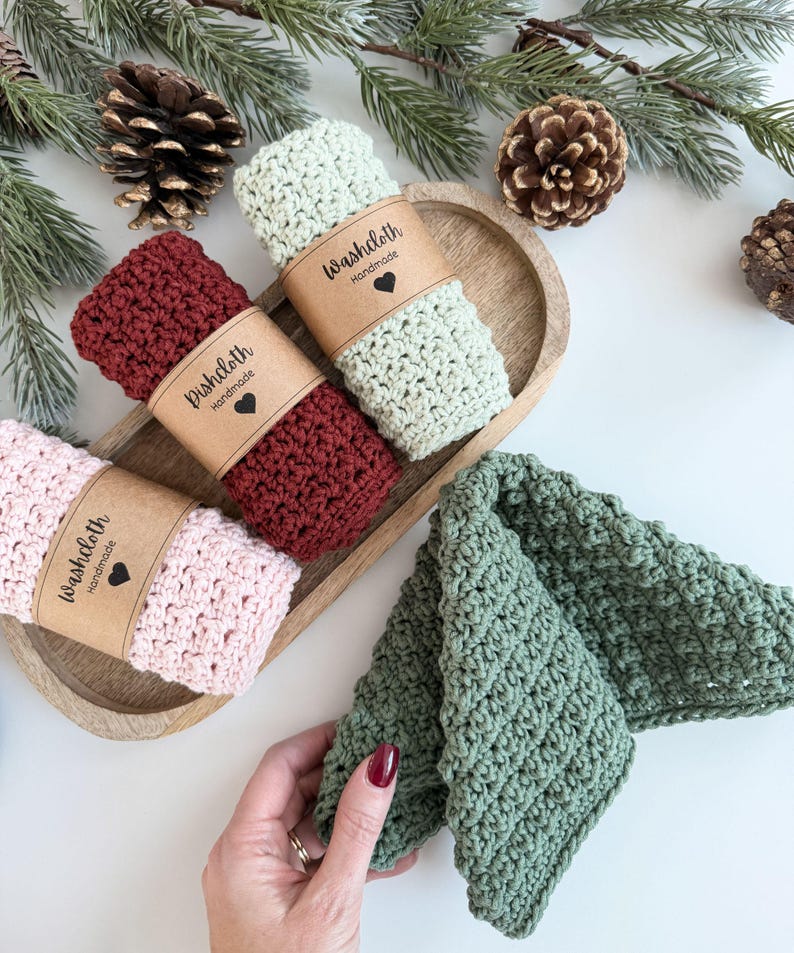May include: A collection of handmade crochet washcloths in various colors, including pink, burgundy, and sage green. Each washcloth is rolled and secured with a brown paper band that reads "Washcloth Handmade" with a heart symbol. The washcloths are displayed on a wooden tray.