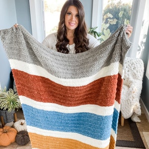 May include: A chunky knit throw blanket featuring horizontal stripes in grey, white, rust orange, white, blue, white, and mustard yellow. The blanket has a thick, textured yarn construction with a visible crochet stitch pattern throughout.
