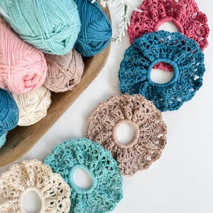 May include: A collection of handmade crochet scrunchies in various colors, including pink, teal, and beige. The scrunchies are displayed alongside balls of yarn in a wooden bowl, creating a craft-focused aesthetic. The scrunchies are a stylish hair accessory.