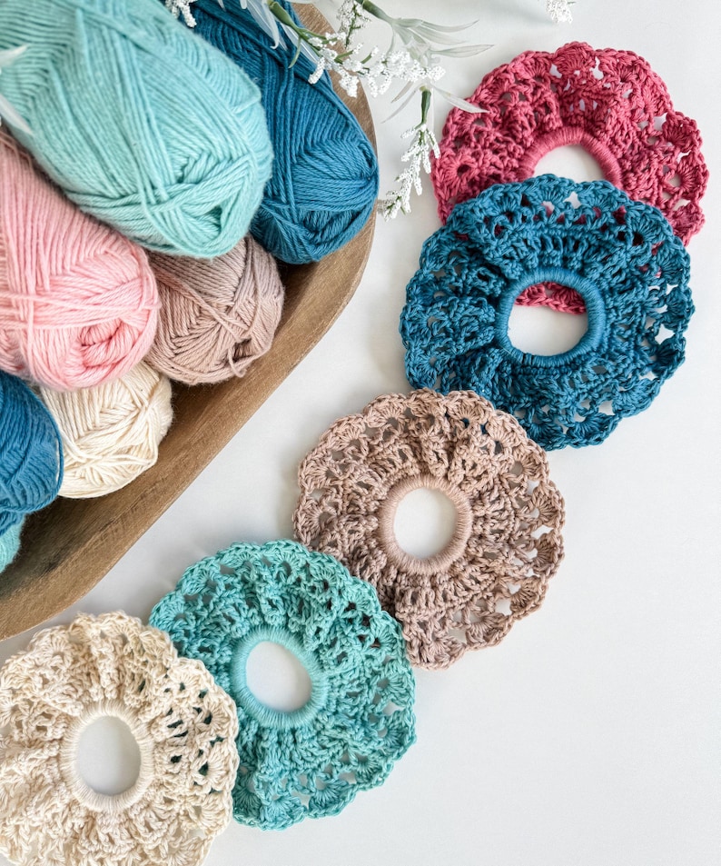May include: A collection of handmade crochet scrunchies in various colors, including pink, teal, and beige. The scrunchies are displayed alongside balls of yarn in a wooden bowl, creating a craft-focused aesthetic. The scrunchies are a stylish hair accessory.