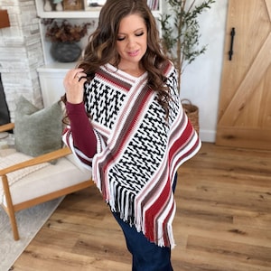 May include: A crochet poncho featuring a geometric pattern in black, white, and maroon. The garment has a V-neckline, horizontal stripes in shades of brown and white, and long fringe trim along the bottom edge, worn over a maroon long-sleeve shirt.