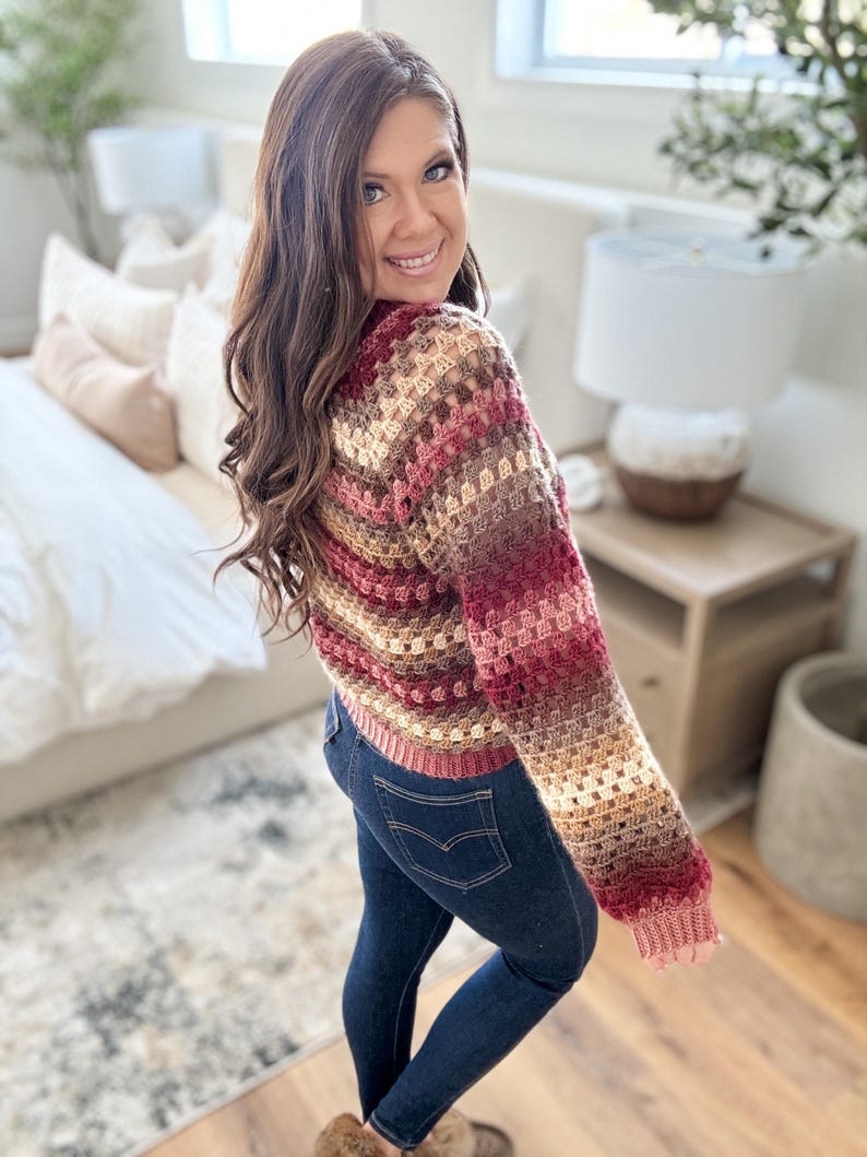Pu&ograve; includere: Un maglione all'uncinetto multicolore nei toni del bordeaux, beige e marrone. Il maglione ha maniche lunghe e orlo e polsini a coste. La persona indossa jeans blu scuro.