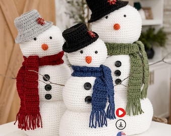 May include: Three crocheted snowmen with black button eyes, orange carrot noses, and black button bodies. Each snowman wears a different colored scarf and hat. The hats are gray, black, and black. The scarves are red, blue, and green.