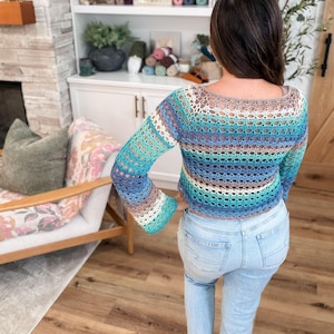 May include: A crochet top with long sleeves in shades of blue, turquoise, brown, and white. The open-weave design reveals the skin beneath. The top is paired with light blue jeans.