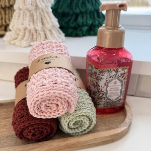 May include: A wooden tray holds rolled washcloths in burgundy, pink, and sage green, each wrapped with a brown paper band. A bottle of pink liquid soap with a floral label and a pump dispenser sits beside the washcloths. Knitted Christmas tree decorations are in the background.