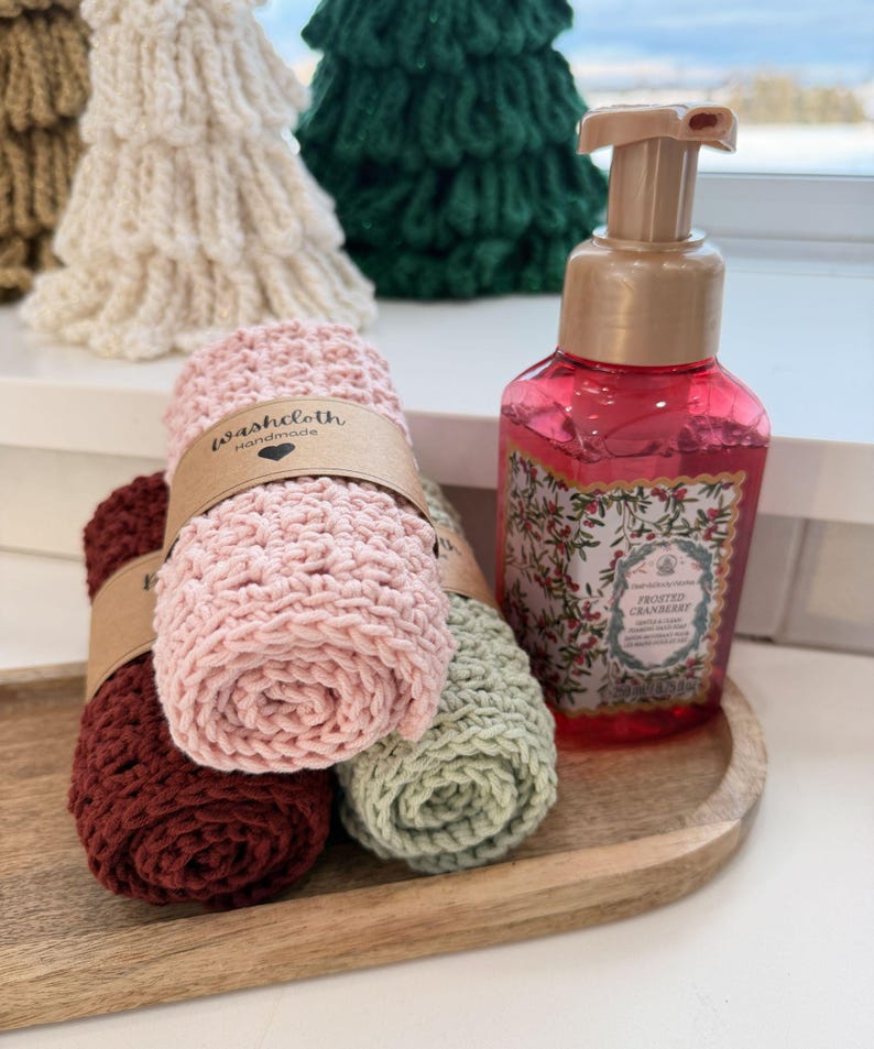 May include: A wooden tray holds rolled washcloths in burgundy, pink, and sage green, each wrapped with a brown paper band. A bottle of pink liquid soap with a floral label and a pump dispenser sits beside the washcloths. Knitted Christmas tree decorations are in the background.