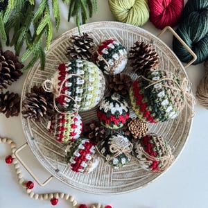 May include: A collection of eight crocheted Christmas ornaments in a variety of green, red, and brown shades. The ornaments are arranged in a white ceramic bowl with pine cones and greenery.