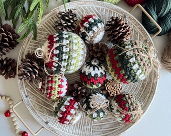 May include: A collection of eight crocheted Christmas baubles in a variety of green, red, and brown shades. The baubles are arranged in a white ceramic bowl with pine cones and greenery.
