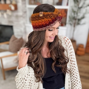 May include: A handmade, crocheted headband in a warm colour palette of orange, brown, and burgundy. The headband features a small leather tag with a logo. The woman in the image is wearing a cream-coloured, open-weave cardigan.