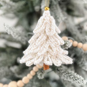 May include: A white crocheted Christmas tree ornament with gold accents. The tree-shaped decoration hangs from a gold string and has a small, brown wooden base. The ornament is displayed on a frosted green Christmas tree.