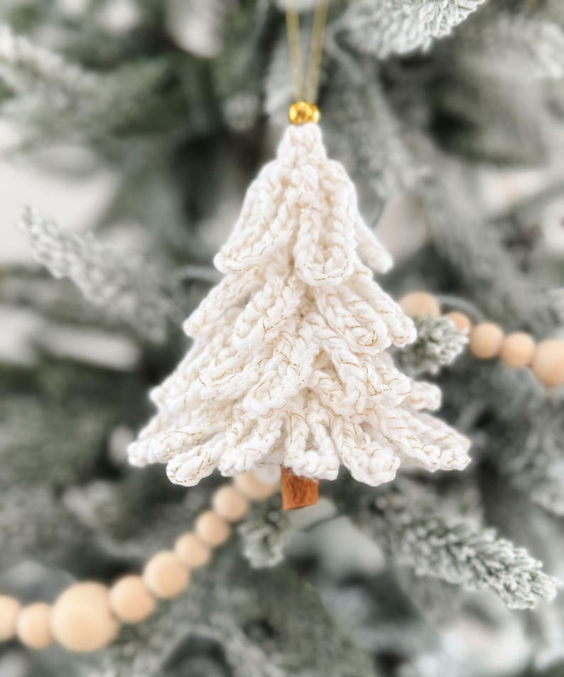 K&ouml;nnte beinhalten: Wei&szlig; geh&auml;kelter Weihnachtsbaumschmuck mit goldenen Akzenten. Der baumf&ouml;rmige Anh&auml;nger h&auml;ngt an einem goldenen Faden und hat einen kleinen, braunen Holzsockel. Der Schmuck befindet sich auf einem gefrosteten gr&uuml;nen Weihnachtsbaum.