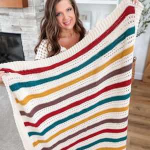 May include: A rectangular crochet blanket featuring a cream-coloured base with horizontal stripes in shades of deep red, teal, mustard yellow, and brown. The textile displays a repeating pattern of solid stitches and openwork sections throughout the design.