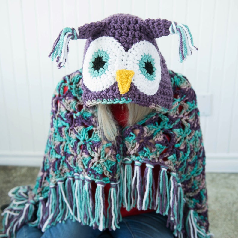 May include: A purple crocheted owl hooded blanket with teal, white, and purple fringe. The owl has white eyes with teal pupils and a yellow beak.