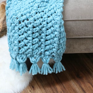 Pu&ograve; includere: Una coperta in crochet blu chiaro con frange. La coperta &egrave; appoggiata su un divano marrone chiaro.