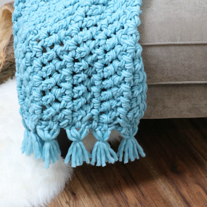 Pu&ograve; includere: Una coperta in crochet blu chiaro con frange. La coperta &egrave; appoggiata su un divano marrone chiaro.