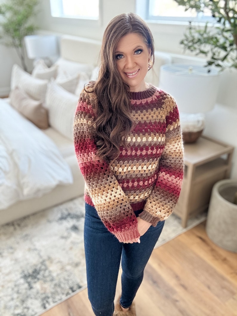 Pu&ograve; includere: Un maglione all'uncinetto nei toni del bordeaux, marrone e beige. Il maglione ha uno scollo tondo e maniche lunghe. La persona nell'immagine indossa jeans blu scuro.