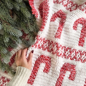 K&ouml;nnte beinhalten: Eine wei&szlig;-rote H&auml;keldecke mit einem Zuckerstangenmuster. Die Decke hat gro&szlig;e rote Zuckerstangenmuster auf wei&szlig;em Hintergrund. Die R&auml;nder sind mit einem rot-wei&szlig;en Muster verziert. Eine Hand ber&uuml;hrt die Decke.