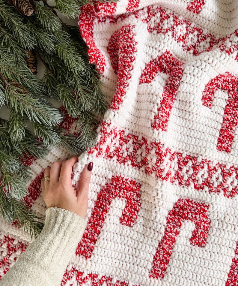 K&ouml;nnte beinhalten: Eine wei&szlig;-rote H&auml;keldecke mit einem Zuckerstangenmuster. Die Decke hat gro&szlig;e rote Zuckerstangenmuster auf wei&szlig;em Hintergrund. Die R&auml;nder sind mit einem rot-wei&szlig;en Muster verziert. Eine Hand ber&uuml;hrt die Decke.