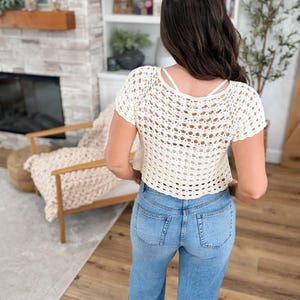 May include: Cream-colored crochet top with short sleeves and a scoop back. The top is paired with light blue denim jeans. The crochet design features an open weave pattern. The model is facing away from the camera.