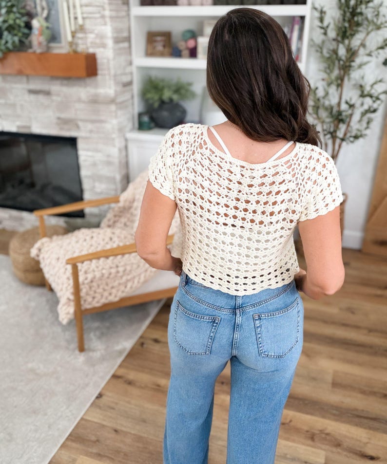 May include: Cream-colored crochet top with short sleeves and a scoop back. The top is paired with light blue denim jeans. The crochet design features an open weave pattern. The model is facing away from the camera.