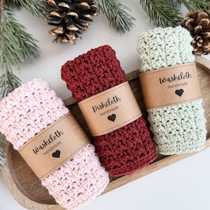 May include: Three handmade crochet washcloths in pink, burgundy, and sage green, each wrapped with a brown paper band. The bands are printed with "Washcloth Handmade" or "Dishcloth Handmade" and a heart. The washcloths are arranged on a wooden tray.