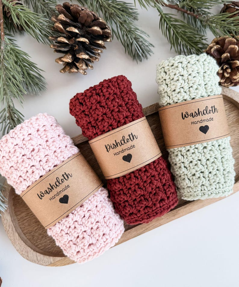 May include: Three handmade crochet washcloths in pink, burgundy, and sage green, each wrapped with a brown paper band. The bands are printed with "Washcloth Handmade" or "Dishcloth Handmade" and a heart. The washcloths are arranged on a wooden tray.