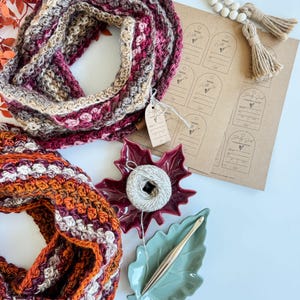 May include: A close-up shot of two crocheted infinity scarves in fall colors. One scarf is primarily burgundy, tan, and pink. The other is orange, burgundy, and white. Also shown are craft supplies: a ball of yarn, a crochet hook, and tags that say "Made With Love".