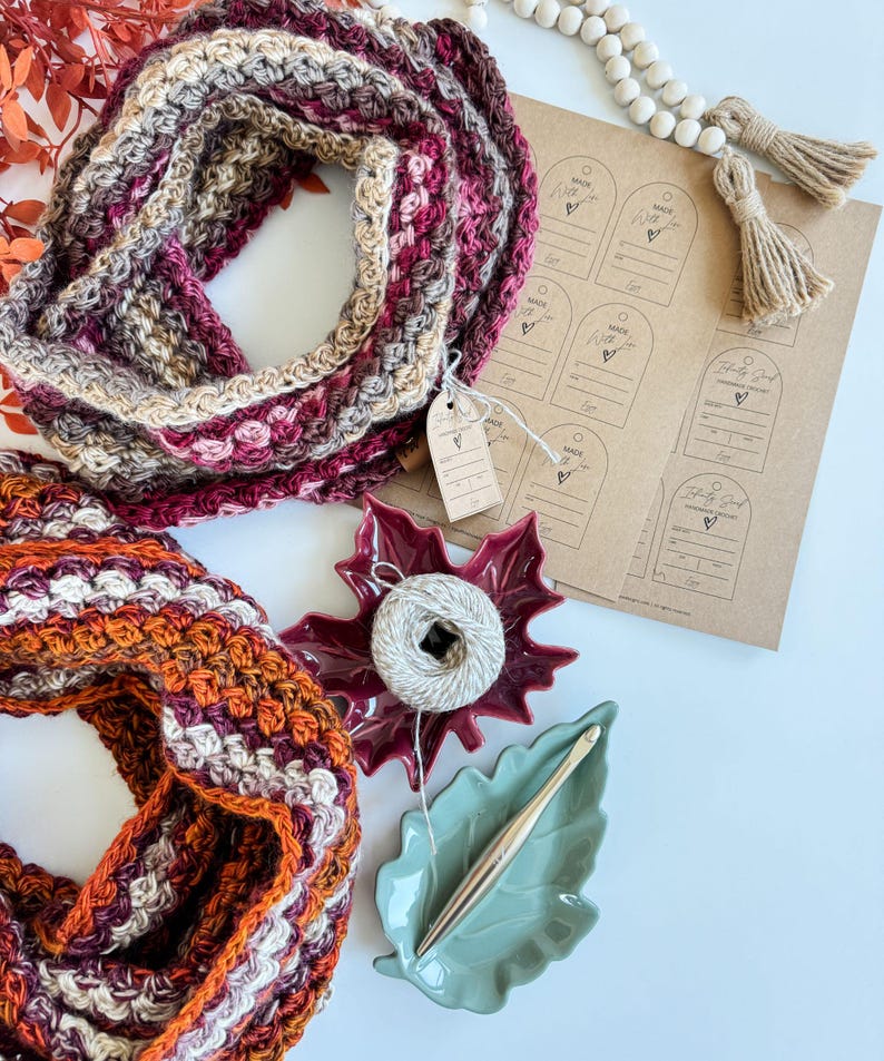 May include: A close-up shot of two crocheted infinity scarves in fall colors. One scarf is primarily burgundy, tan, and pink. The other is orange, burgundy, and white. Also shown are craft supplies: a ball of yarn, a crochet hook, and tags that say "Made With Love".