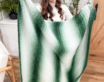 Può includere: Una coperta rettangolare all'uncinetto con un motivo sfumato che passa dal verde bosco scuro al verde salvia chiaro e al bianco. Il tessuto presenta un motivo a punti ripetitivo e testurizzato, creando un aspetto morbido e stratificato.