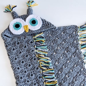 May include: Gray crocheted owl hooded blanket with colorful fringe. The owl has large white eyes with teal and black accents and a yellow beak.