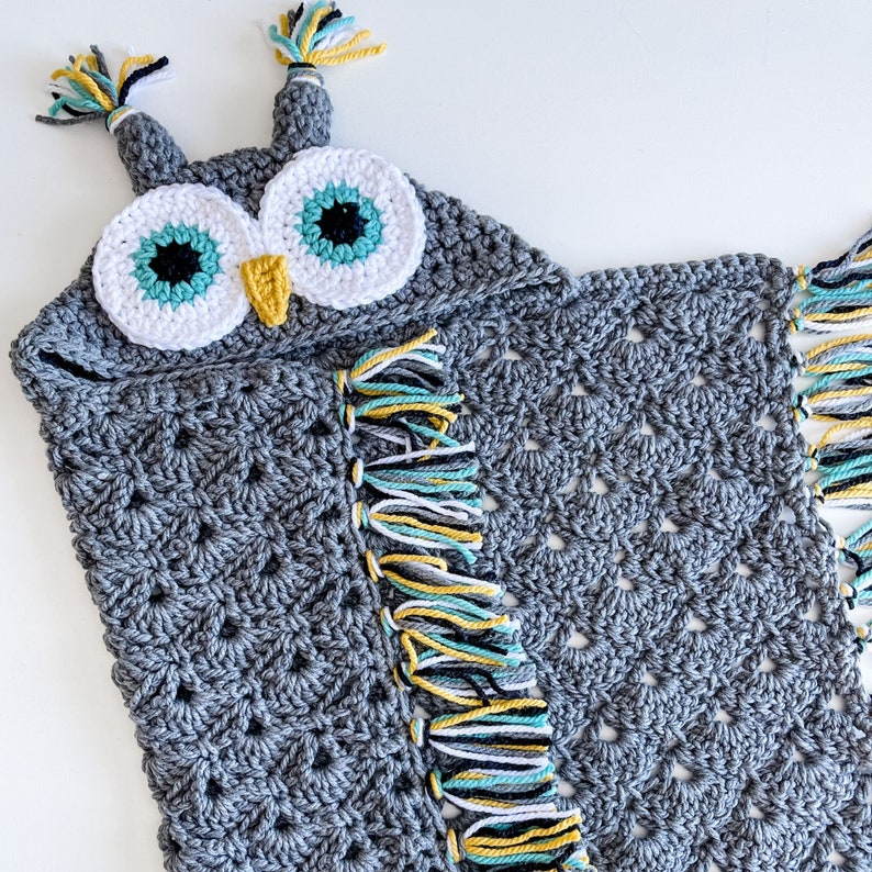 May include: Gray crocheted owl hooded blanket with colorful fringe. The owl has large white eyes with teal and black accents and a yellow beak.