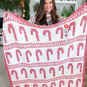 May include: A white and red crocheted blanket featuring a repeating candy cane pattern. The blanket has a red border and candy cane designs. The blanket is held up by a person.