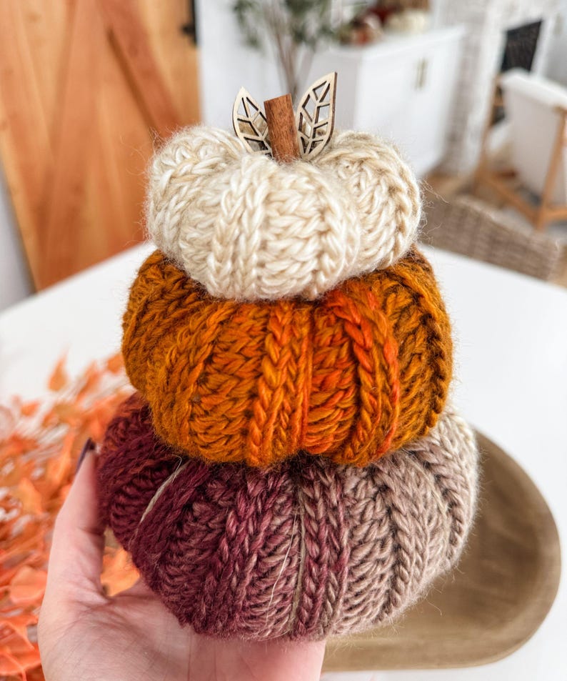 K&ouml;nnte beinhalten: Ein Stapel aus drei gestrickten K&uuml;rbissen in Herbstfarben. Der oberste K&uuml;rbis ist cremefarben, der mittlere orangefarben und der untere weinrot und braun. Ein Holzstiel und Bl&auml;tter akzentuieren den obersten K&uuml;rbis.