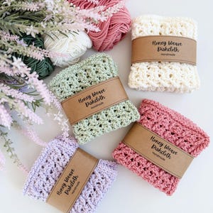Puede incluir: Una colecci&oacute;n de pa&ntilde;os de cocina Honey Weave hechos a mano en varios colores: crema, verde claro, rosa y lavanda. Cada pa&ntilde;o est&aacute; envuelto con una banda de papel marr&oacute;n que dice "Honey Weave Dishcloth Handmade". Hilo en tonos rosa, blanco y verde en el fondo.