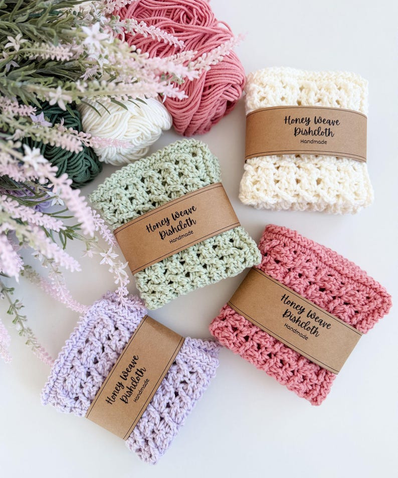 Puede incluir: Una colecci&oacute;n de pa&ntilde;os de cocina Honey Weave hechos a mano en varios colores: crema, verde claro, rosa y lavanda. Cada pa&ntilde;o est&aacute; envuelto con una banda de papel marr&oacute;n que dice "Honey Weave Dishcloth Handmade". Hilo en tonos rosa, blanco y verde en el fondo.