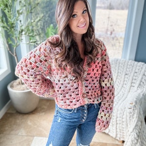 May include: A pink, white, and beige crochet cardigan with a button closure. The cardigan is cropped and has a loose fit. The model is wearing blue jeans.