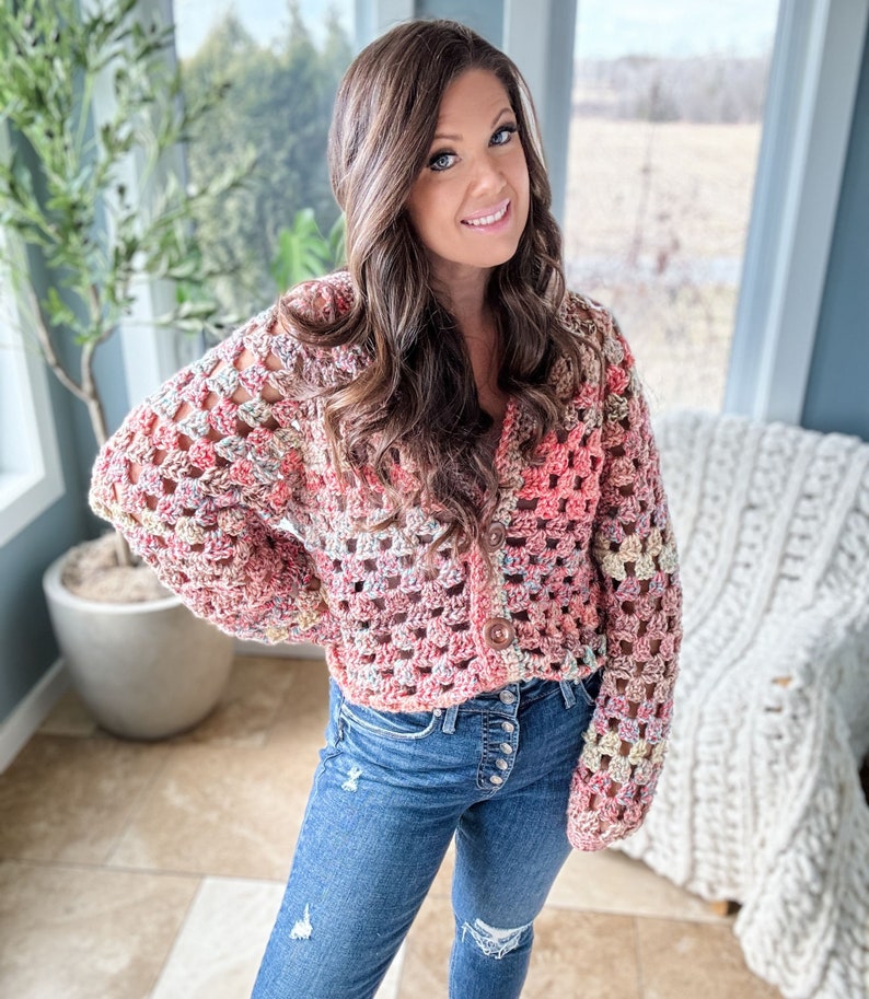 May include: A pink, white, and beige crochet cardigan with a button closure. The cardigan is cropped and has a loose fit. The model is wearing blue jeans.