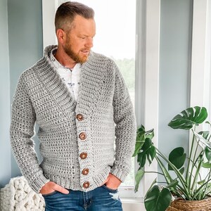 May include: A grey crocheted cardigan with a v-neck and button closure. The cardigan is worn over a white button-down shirt and blue jeans.