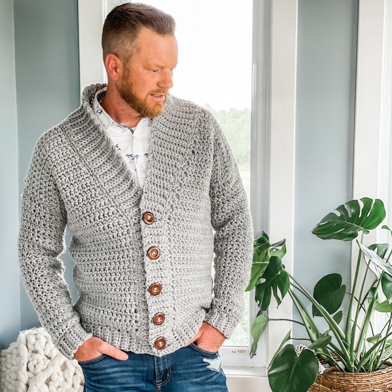 May include: A grey crocheted cardigan with a v-neck and button closure. The cardigan is worn over a white button-down shirt and blue jeans.