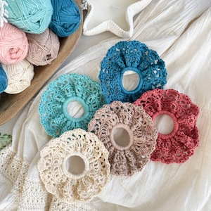 May include: A collection of five handmade crochet scrunchies in various colors: teal, light blue, beige, mauve, and coral. The scrunchies are arranged on a white fabric surface, with balls of yarn in a wooden bowl in the background.