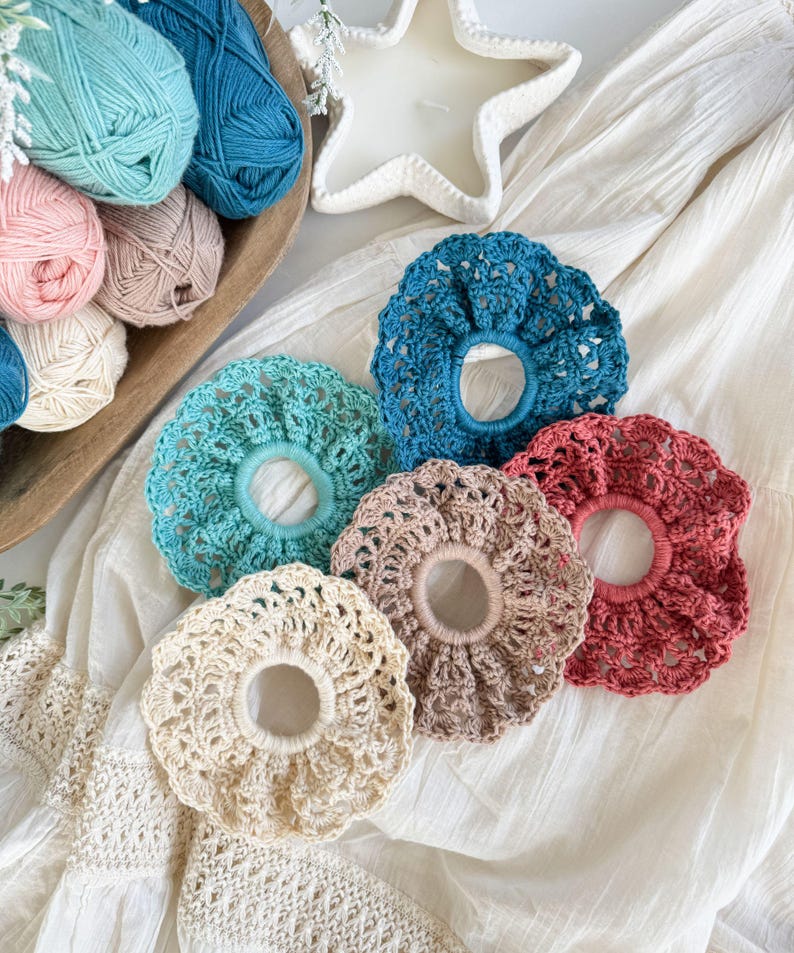 May include: A collection of five handmade crochet scrunchies in various colors: teal, light blue, beige, mauve, and coral. The scrunchies are arranged on a white fabric surface, with balls of yarn in a wooden bowl in the background.
