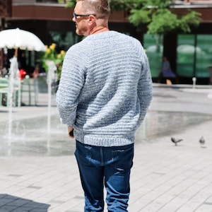 May include: A light blue sweater with a ribbed knit design. The sweater is paired with dark blue jeans. The person is wearing sunglasses and walking outdoors on a sunny day.